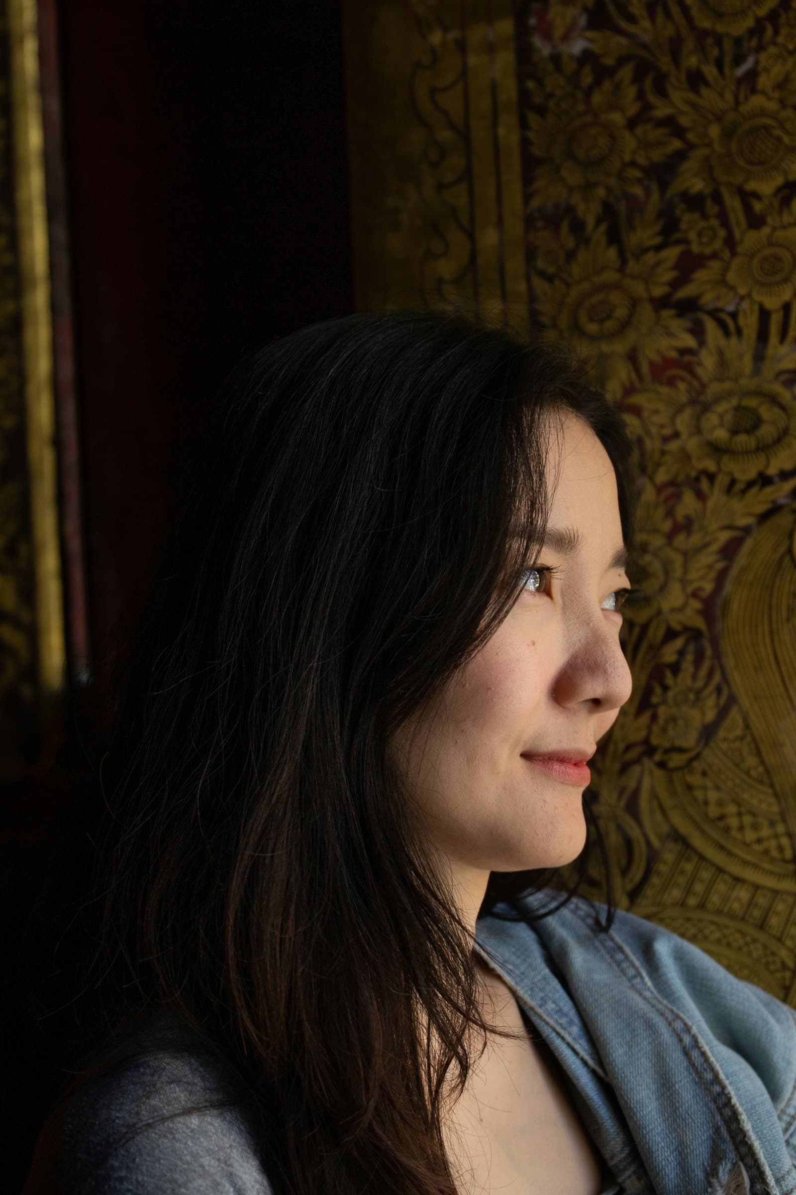 A woman with long dark hair smiles softly as she looks across, lit by warm low light